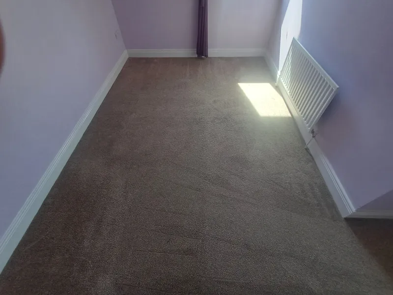 An empty bedroom with a freshly cleaned grey carpet and a radiator under the window